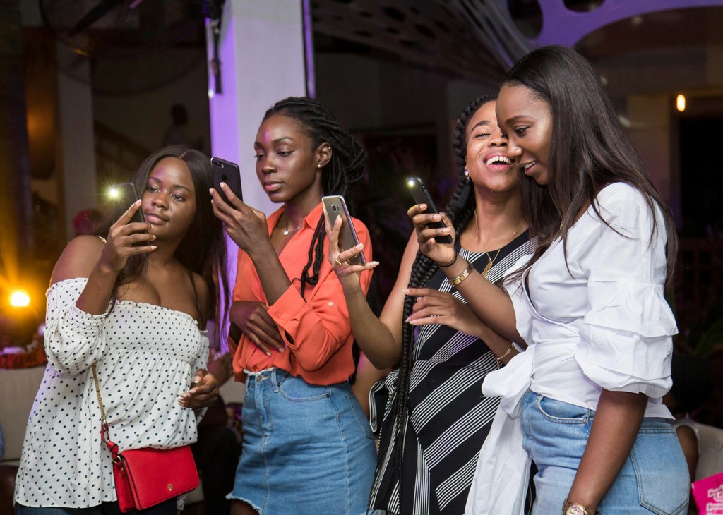  A group of four young women enjoying a night out, capturing moments with their smartphones, smiling and laughing, dressed in stylish casual outfits with a lively indoor ambiance.