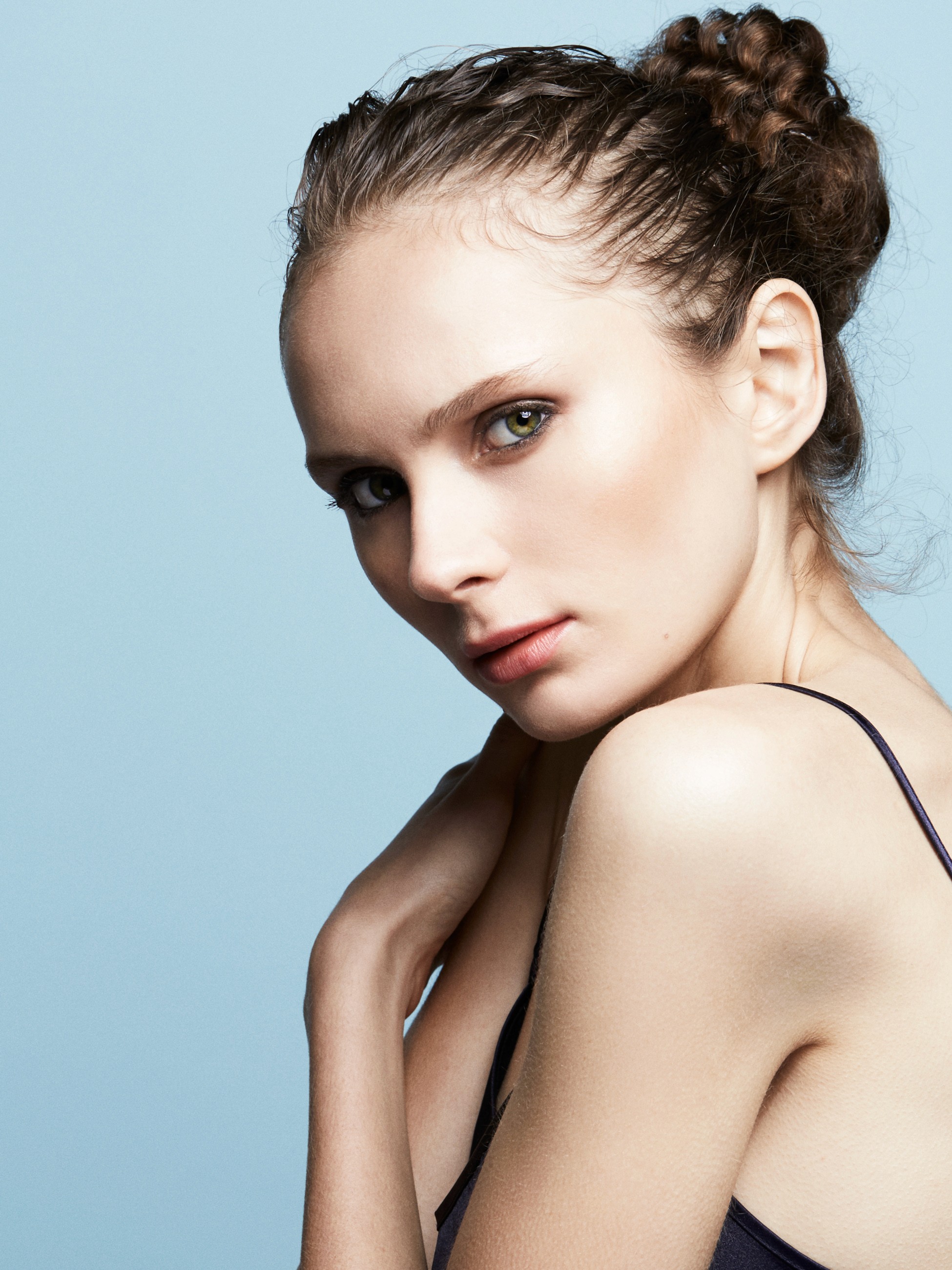 A female model with her curly brunette hair tide back wearing a dark blue string strapped singlet sitting sideways with her chin on her shoulder on an even light blue background.