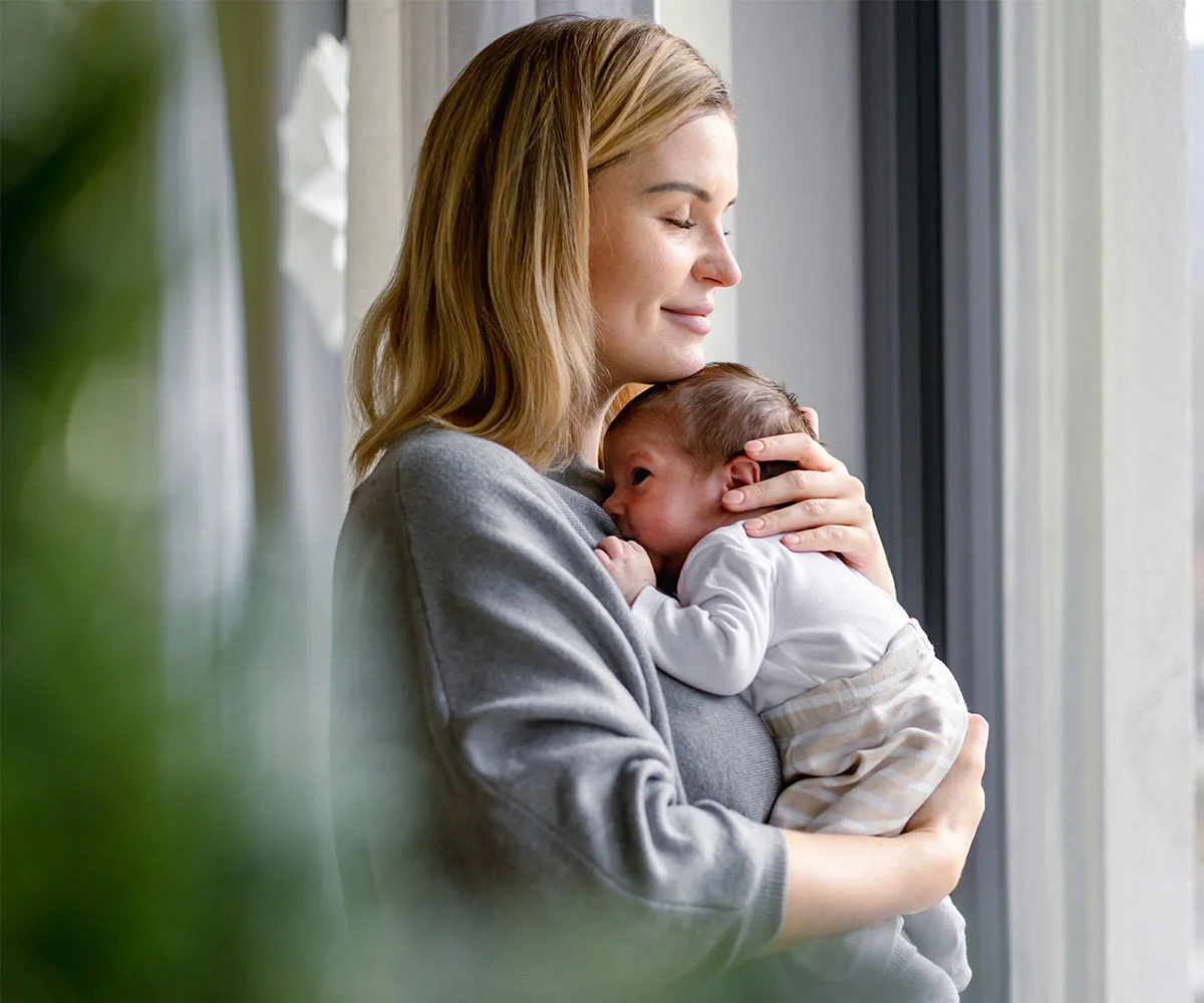 Mother holding her newborn child near a window, representing compassionate and family-centered patient care.