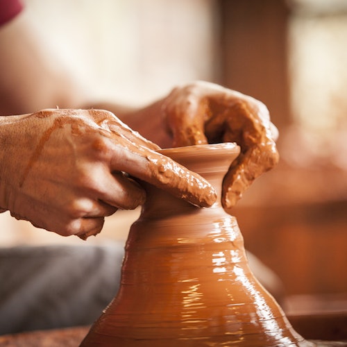Close-up of hands shaping wet clay on a pottery wheel, forming a vase against a soft-focus background.