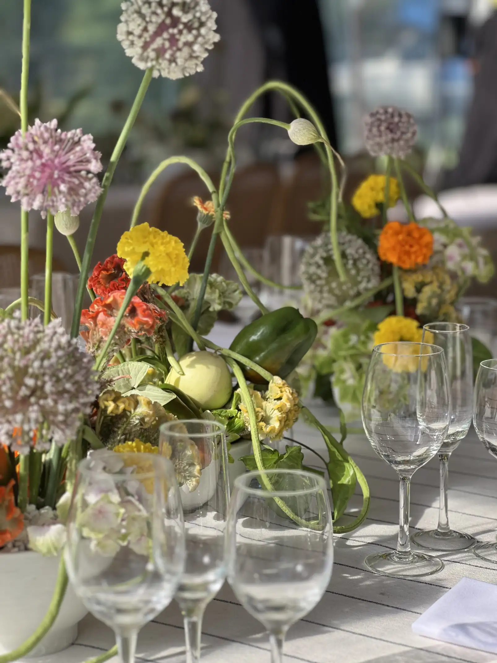 Centro de mesa contemporáneo con flores en tonos cálidos y verdes, dispuesto entre copas de cristal y luz natural.