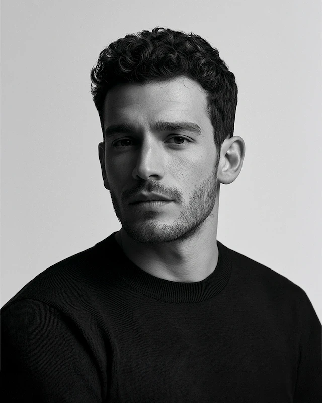 Black-and-white studio portrait of a man with short curly hair and light stubble, wearing a dark crewneck sweater.