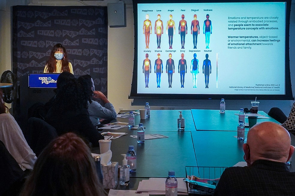 A woman dressed in yellow is giving a speech, and she has the audience sitting around the table and next to her. On the right side of the wall is an image projected representing different body shapes with explanatory text nest to each illustration