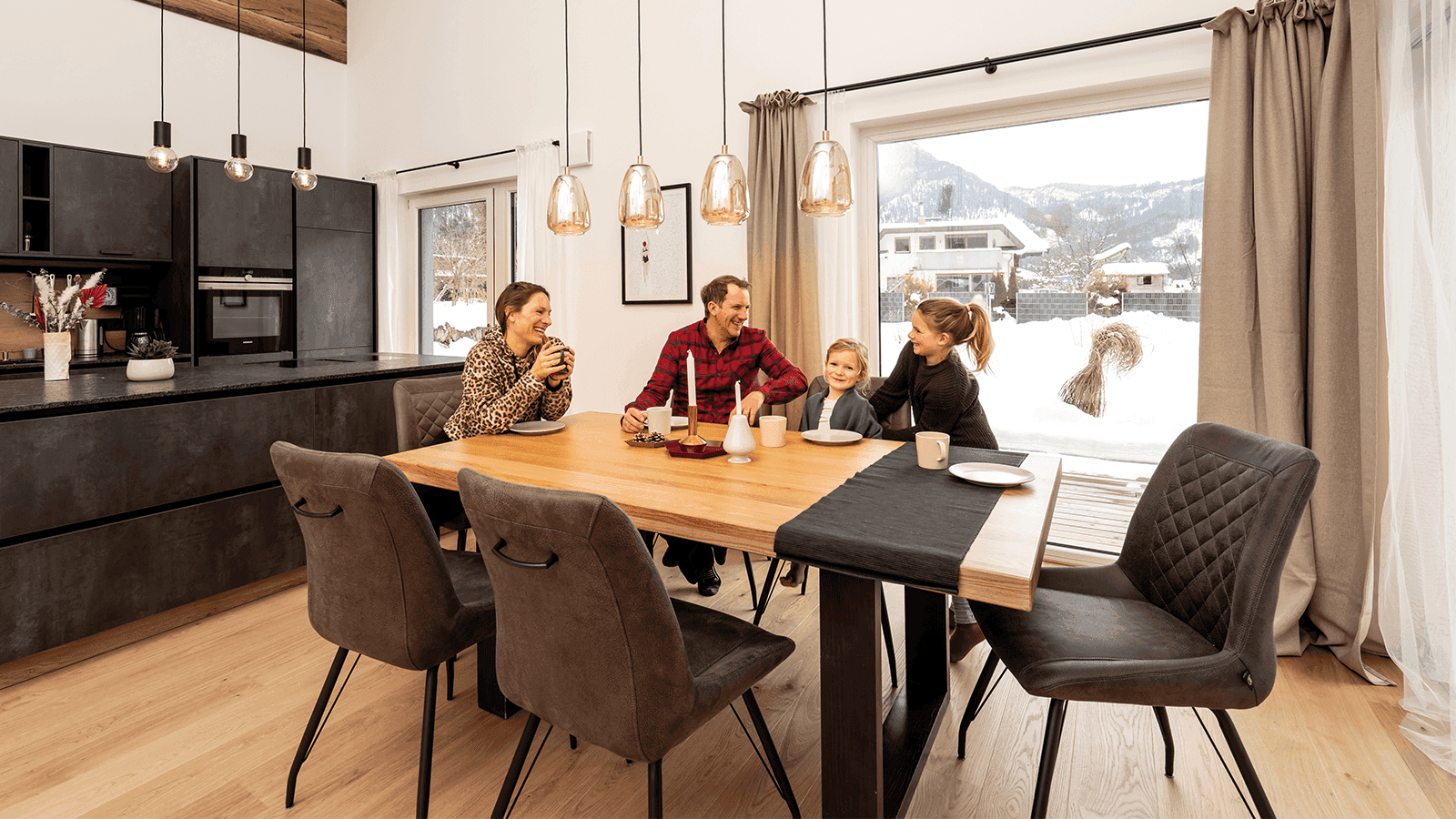 Eine Familie sitzt an einem Holztisch in einem hellen Essbereich mit grosser Fensterfront auf verschneite Berge und genießt eine Mahlzeit.