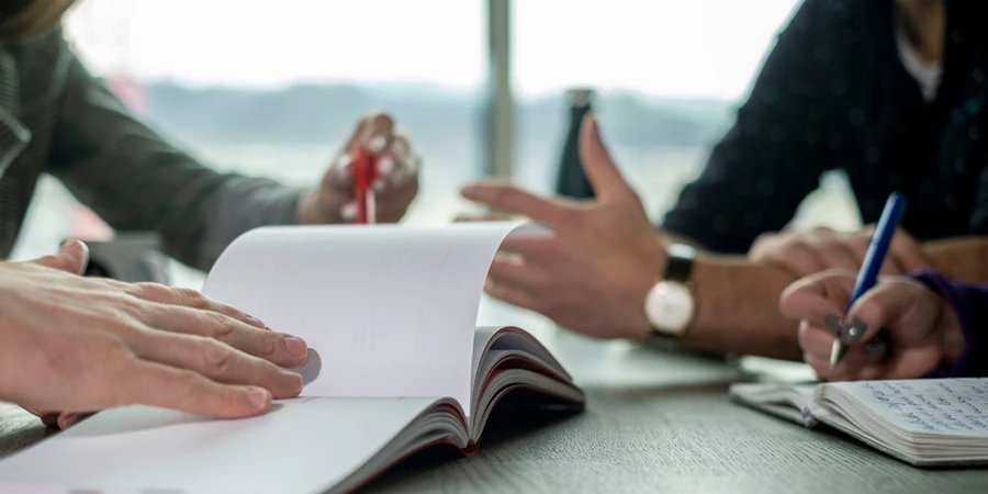Personen sitzen an einem Tisch und diskutieren Ideen mit offenen Notizbüchern und Stiften während eines gemeinsamen Meetings.