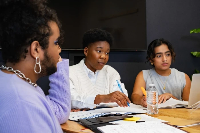 A imagem mostra três pessoas de diferentes gêneros participando de uma reunião de trabalho ou sessão de estudo. Elas estão sentadas ao redor de uma mesa, com cadernos, papéis, canetas e uma garrafa de água visíveis. A pessoa no centro parece estar falando ou explicando algo, enquanto as outras duas estão prestando atenção e tomando notas. O ambiente é descontraído, com roupas casuais, e parece um espaço colaborativo de trabalho ou estudo. A cena transmite diversidade e inclusão, destacando a interação entre diferentes indivíduos em um ambiente de diálogo e troca de ideias.