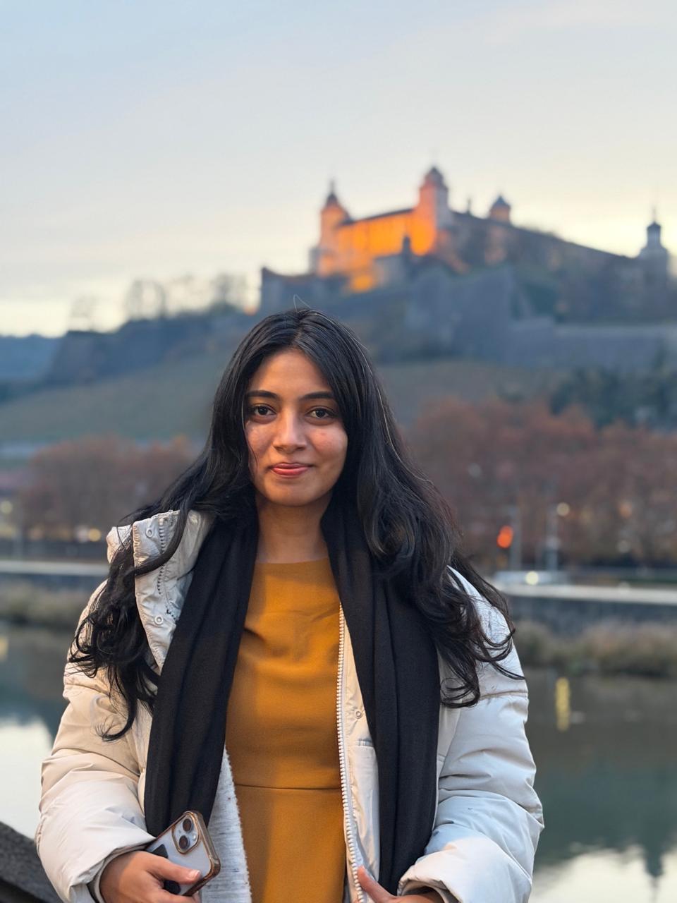 A smiling woman with her arms crossed, standing against a dark green background. She has long, dark hair.