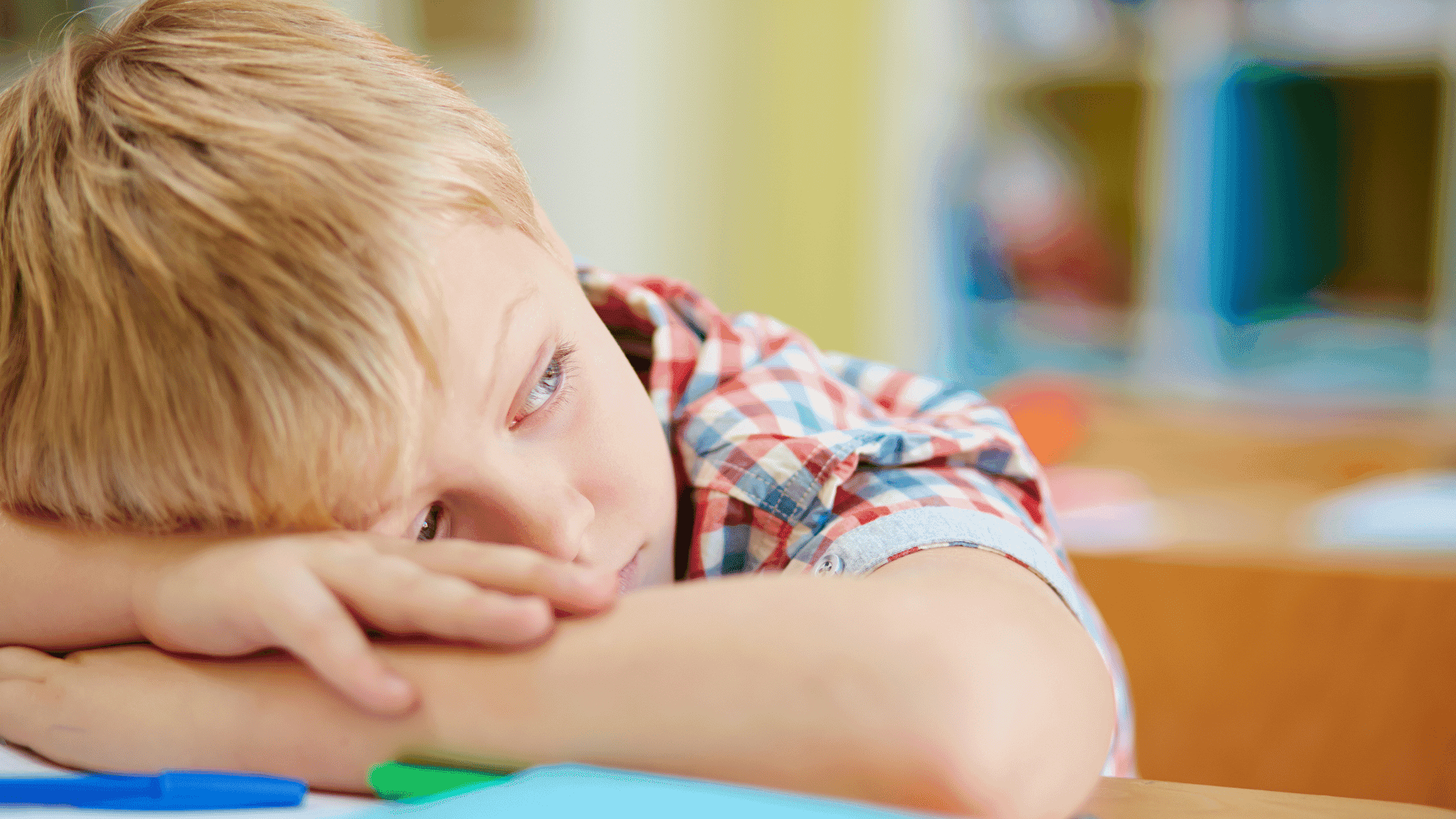 A boy sitting at a table with his head in his arms, looking tired and fed up.