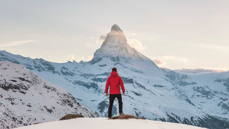 Person is standing on a snowy mountain and looking towards the peak of another mountain.