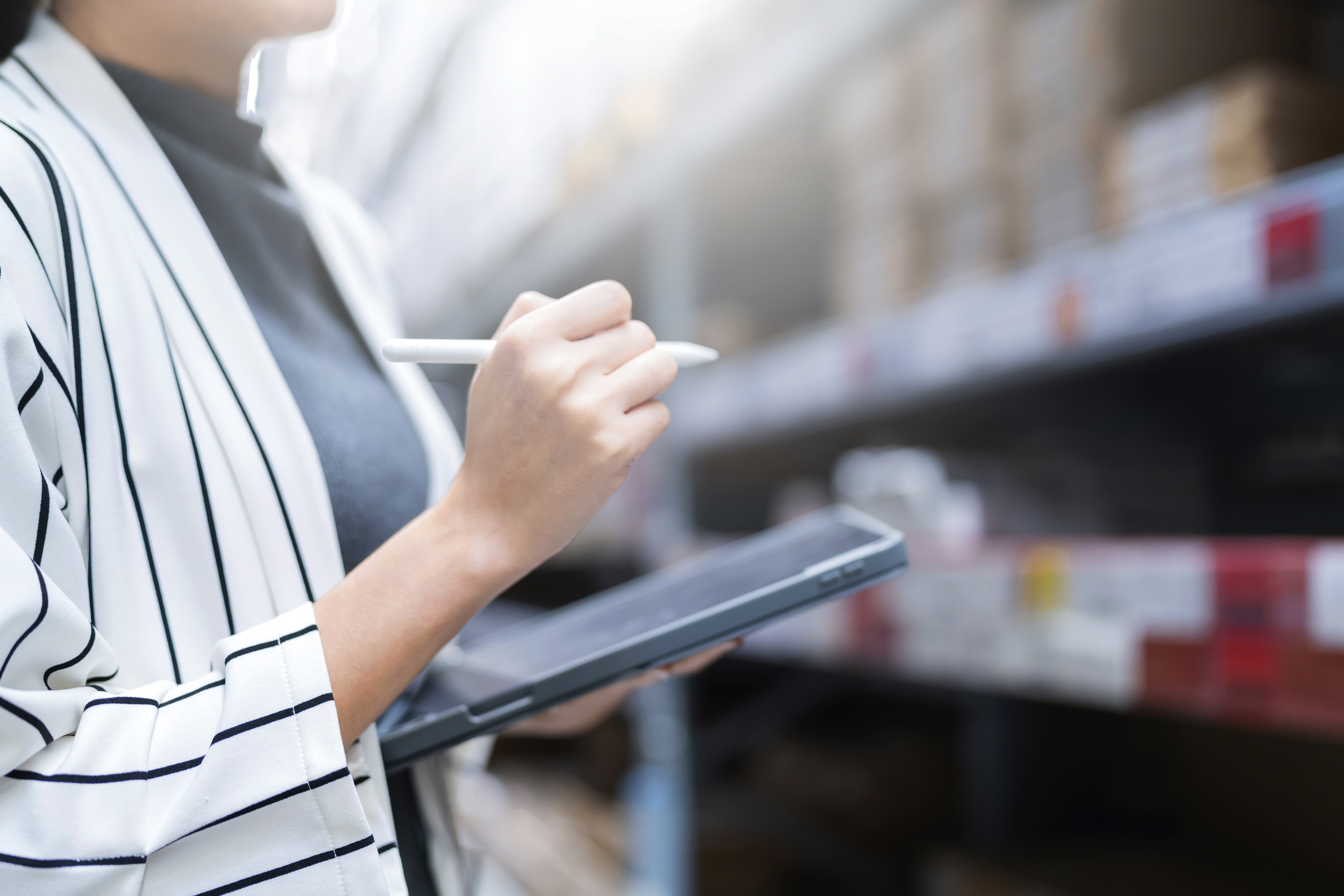 A person in a striped blazer uses a tablet with a stylus in a warehouse setting. Shelves filled with boxes are blurred in the background, emphasising focus and efficiency.