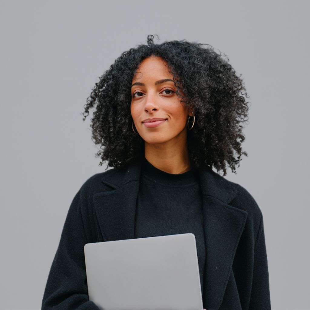 Portrait of a smiling woman with curly hair, wearing a black coat and holding a laptop.