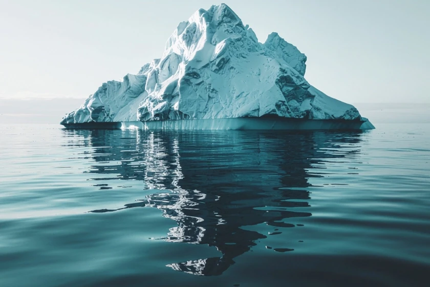 A large iceberg floats in a calm sea, with its jagged white surface illuminated by cool light; beneath the water, the darker and more massive submerged portion is visible, reflected in the still waters.