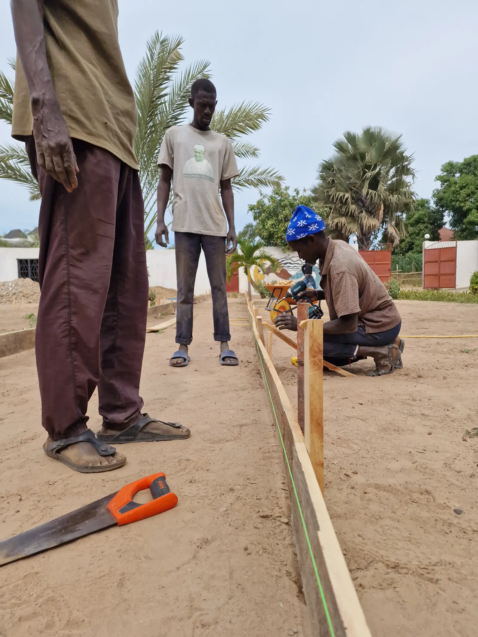 Two workers in a construction site measure and prepare the groundwork under a sunny sky with palm trees nearby.