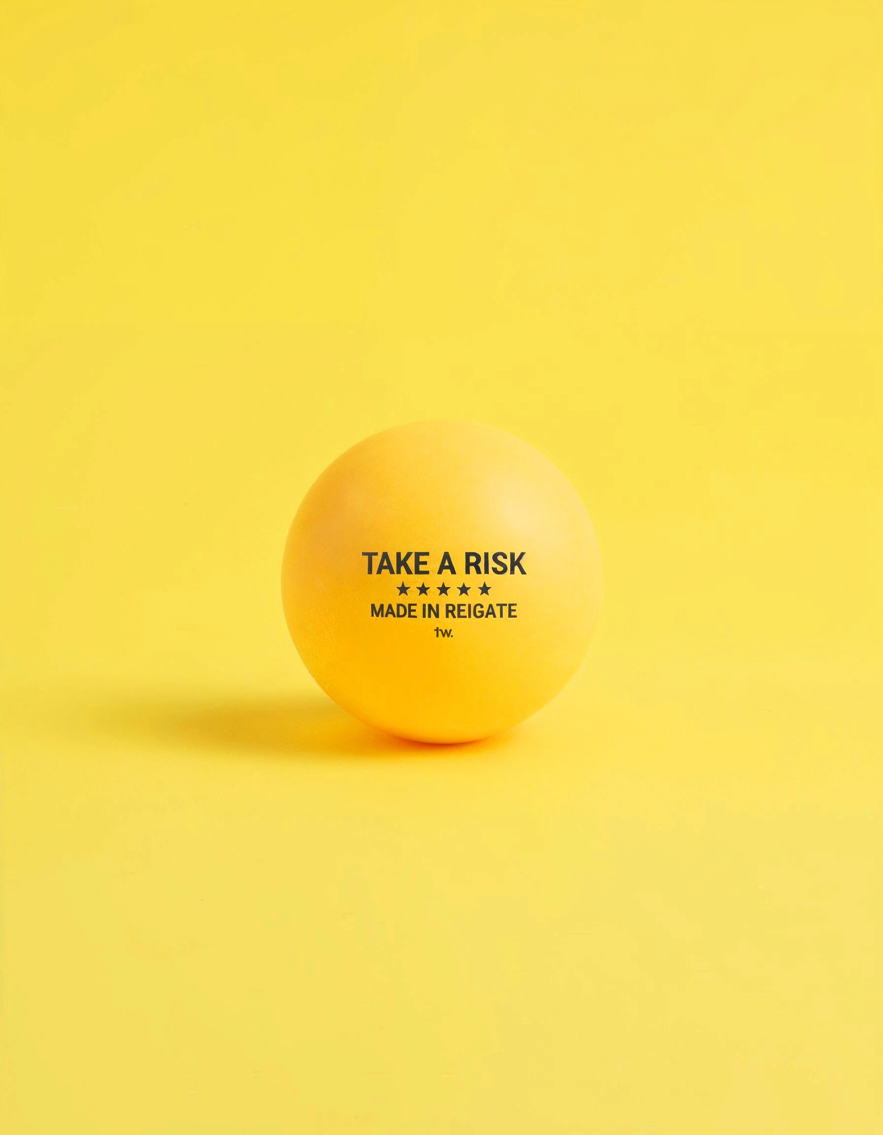 A yellow ping pong ball on a yellow background with TAKE A RISK printed on it