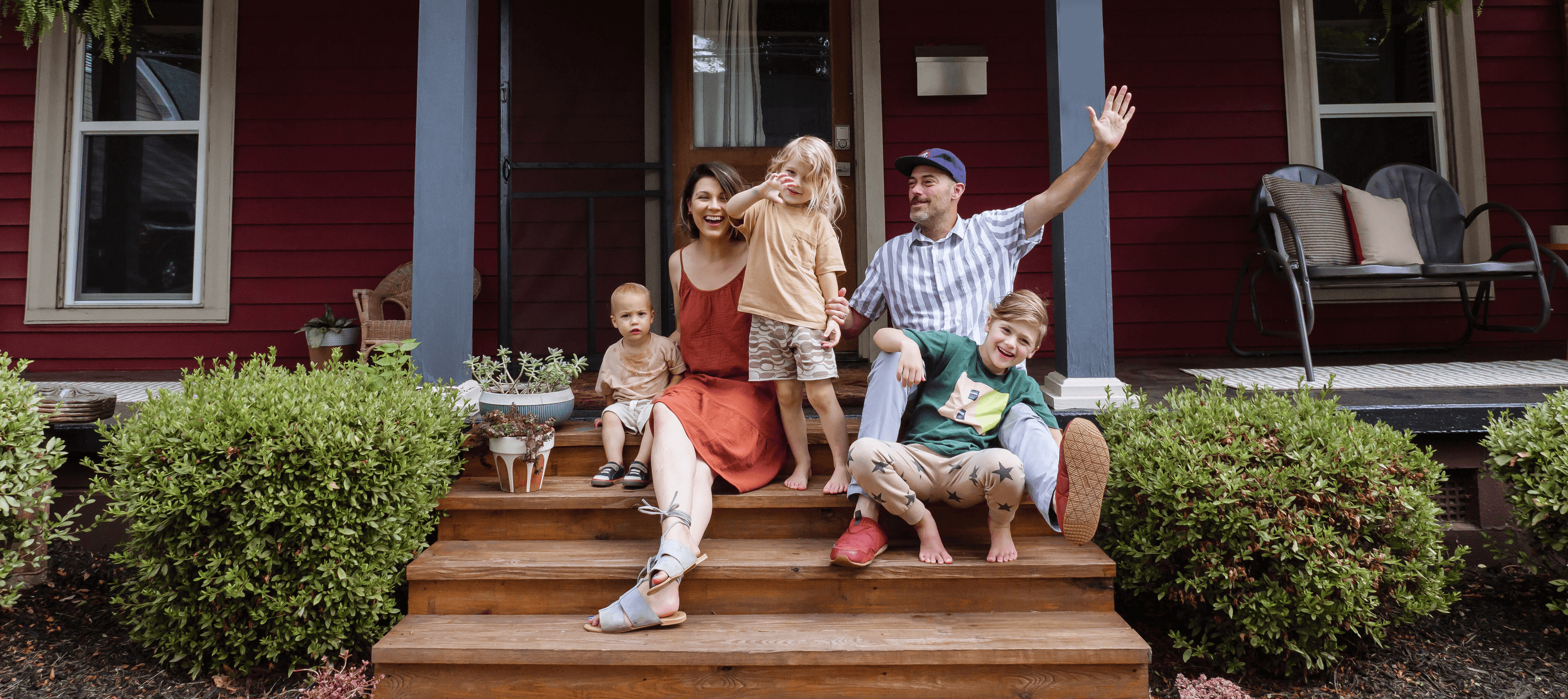 A family with 2 parents and 3 children celebrating on the front porch of a red house.