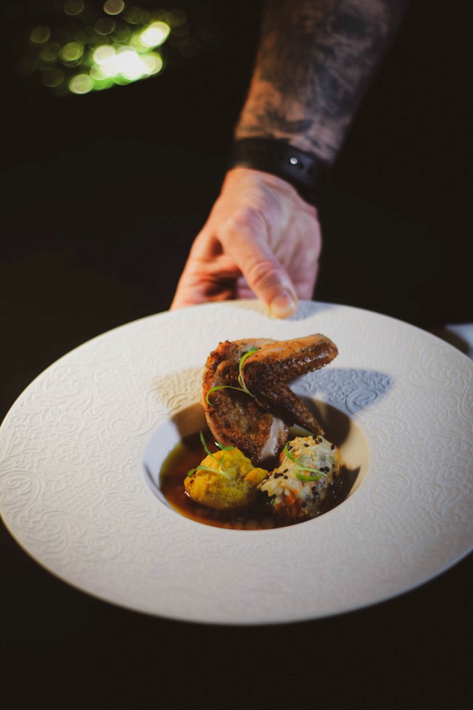 A chef presents a plated dish of roasted game hen with a side of rice and vegetables.