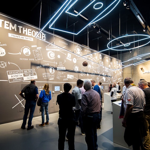 人們在一個現代展覽空間觀看牆壁，上面有圖表和文字，並由天花板上安裝的霓虹藍燈照亮。