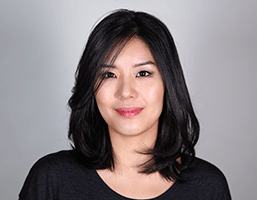 The image shows a woman with shoulder-length black hair styled with bangs that fall across her forehead. She is wearing a black top and facing the camera directly against a plain gray background. Her expression is neutral, and she is looking directly at the camera. The lighting in the image is soft and even. No other objects or text are visible in the image.