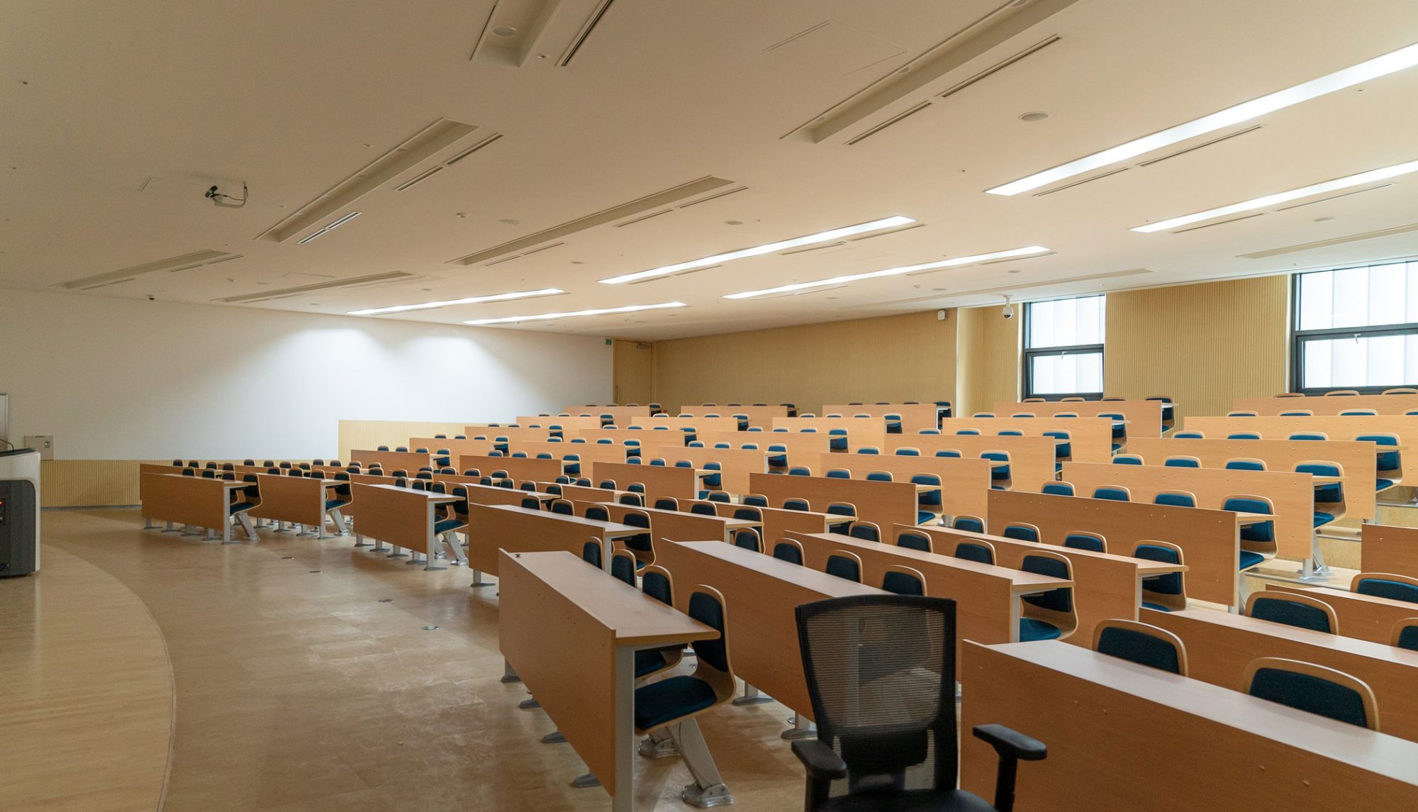 A spacious classroom with rows of desks and chairs, bright lighting, and a few students seated.