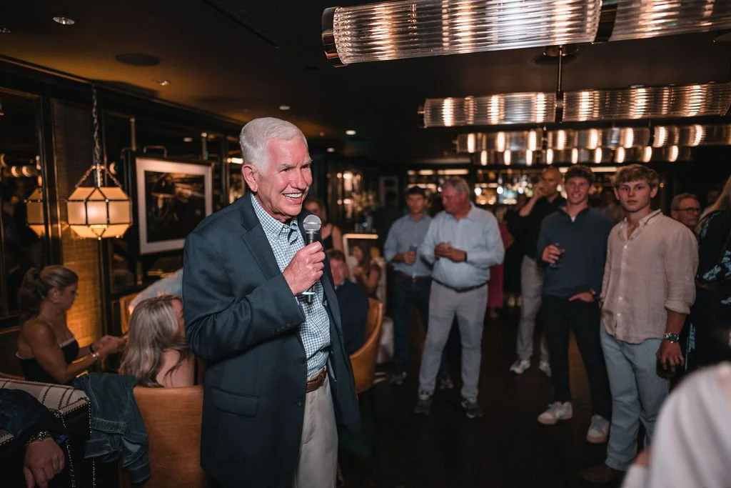 A smiling man holds a microphone while wedding guests gather around him in a lounge.