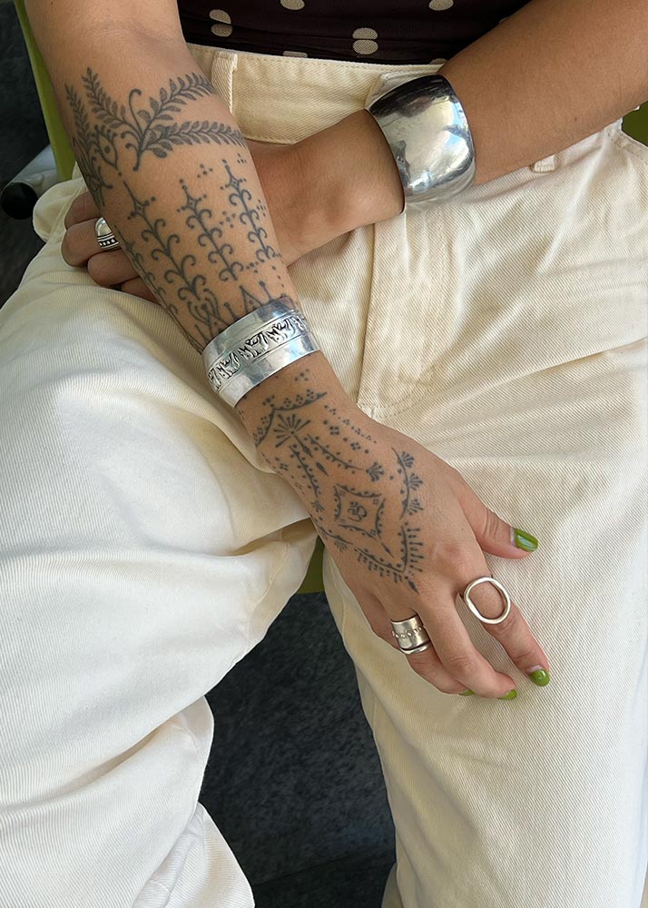 Person with tattoos on arm and hand wearing silver rings and bracelets, sitting on a white surface.