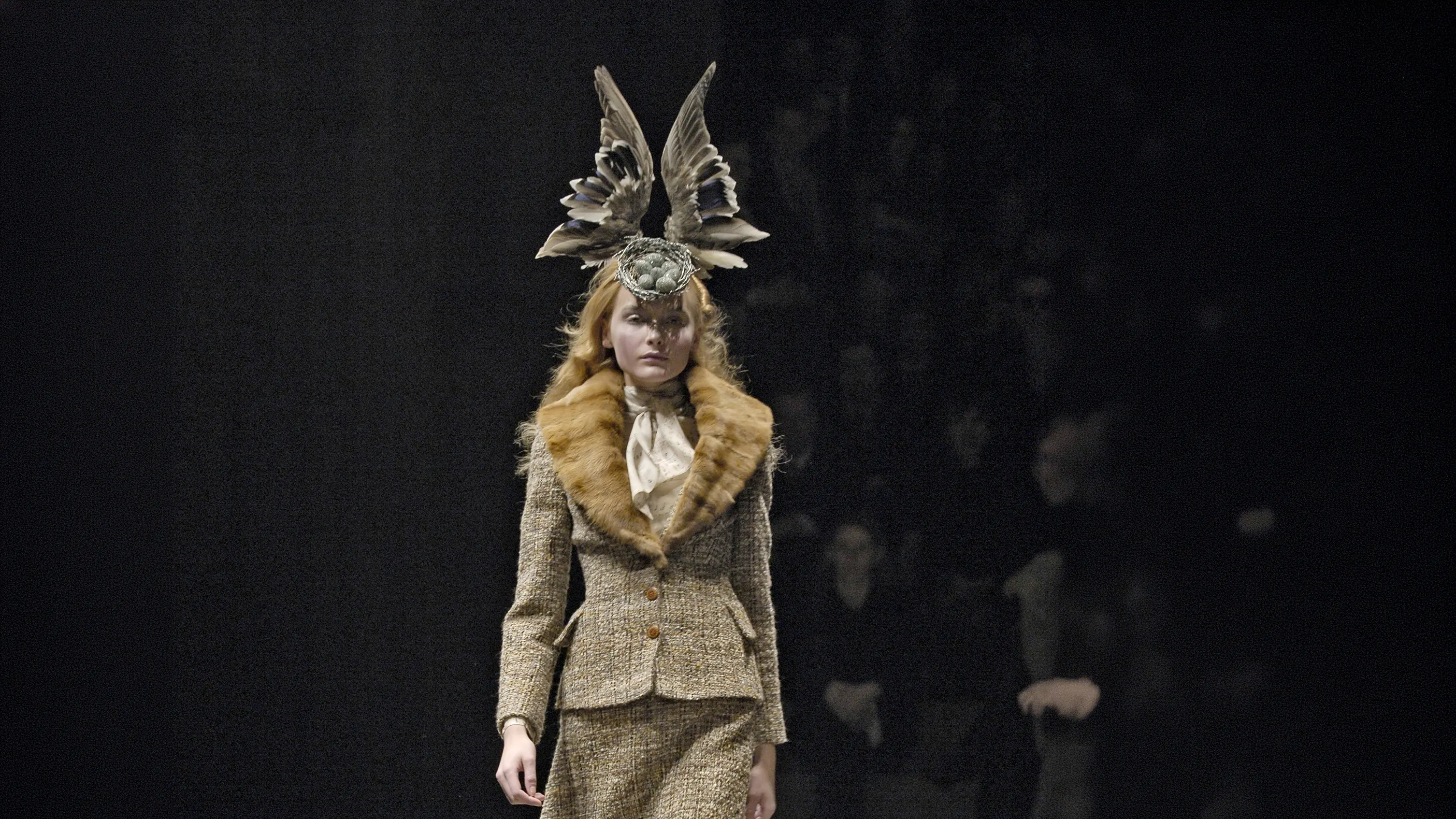 Model on a dark runway wearing a Shaun Leane avant-garde bird's nest headpiece featuring two realistic upright bird wings and small blue eggs, paired with a tweed jacket and fur collar.