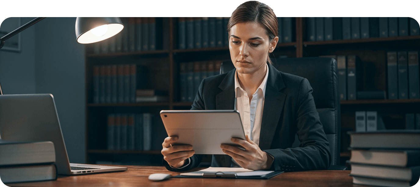 A woman in a suit examines documents while sitting at a desk in a well-lit office with bookshelves.