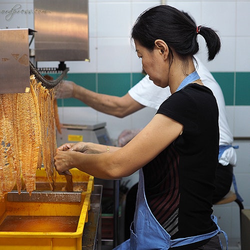 A woman in a black shirt and blue apron processes strips of dough with a machine in a tiled kitchen.