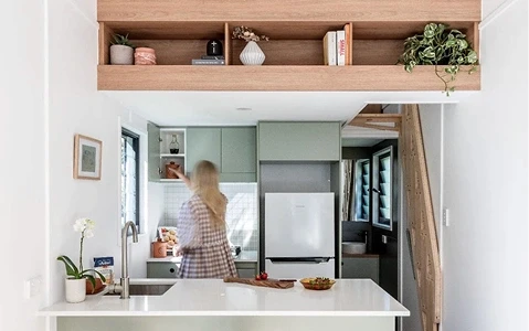 A woman reaching into a sage green cabinet in a modern, minimalist tiny house kitchen featuring white countertops and a wood-toned loft shelf.
