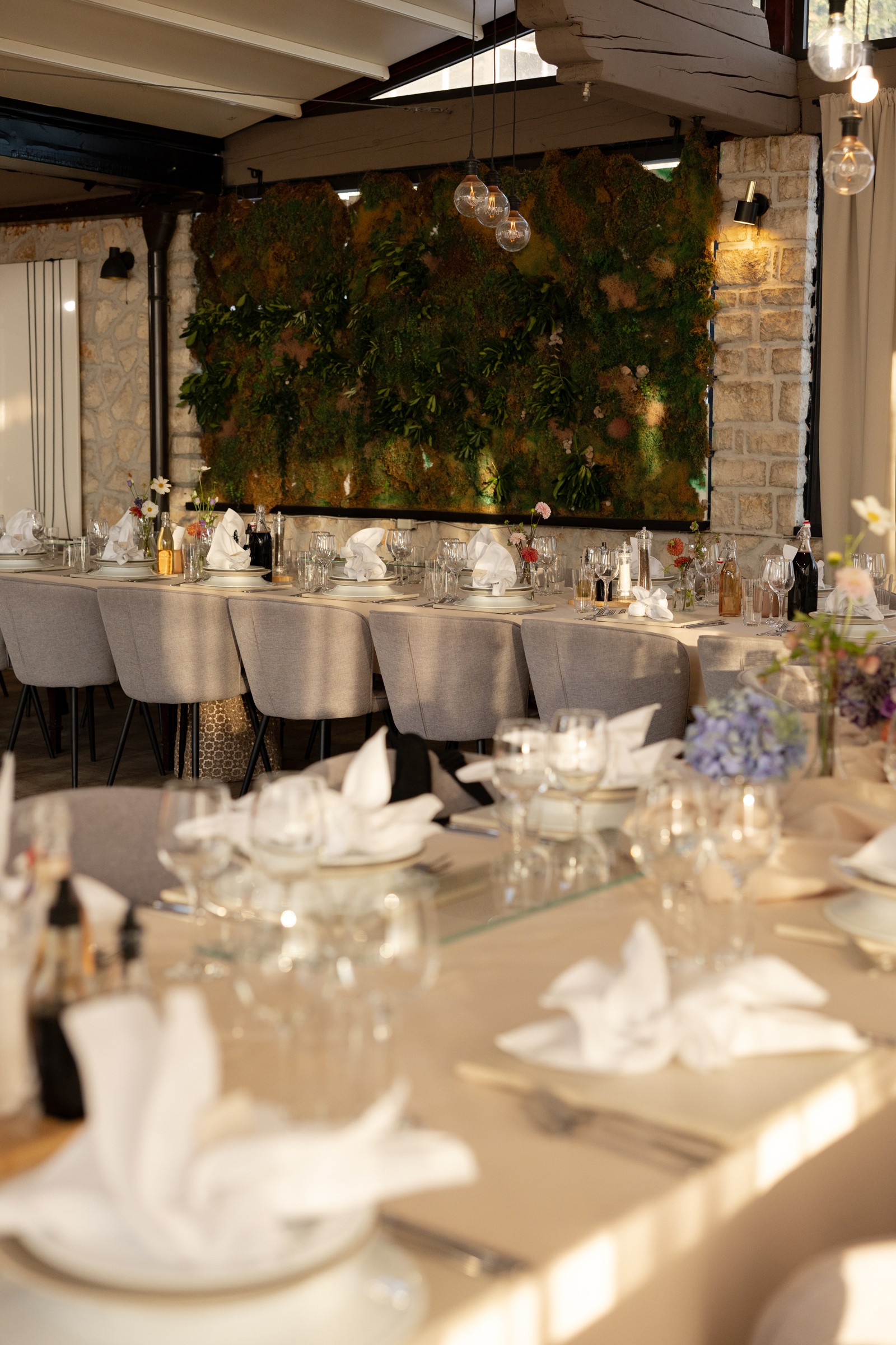 Dining room in a restaurant with a long table, set with plates, glasses, and flower arrangements, with a green plant wall behind