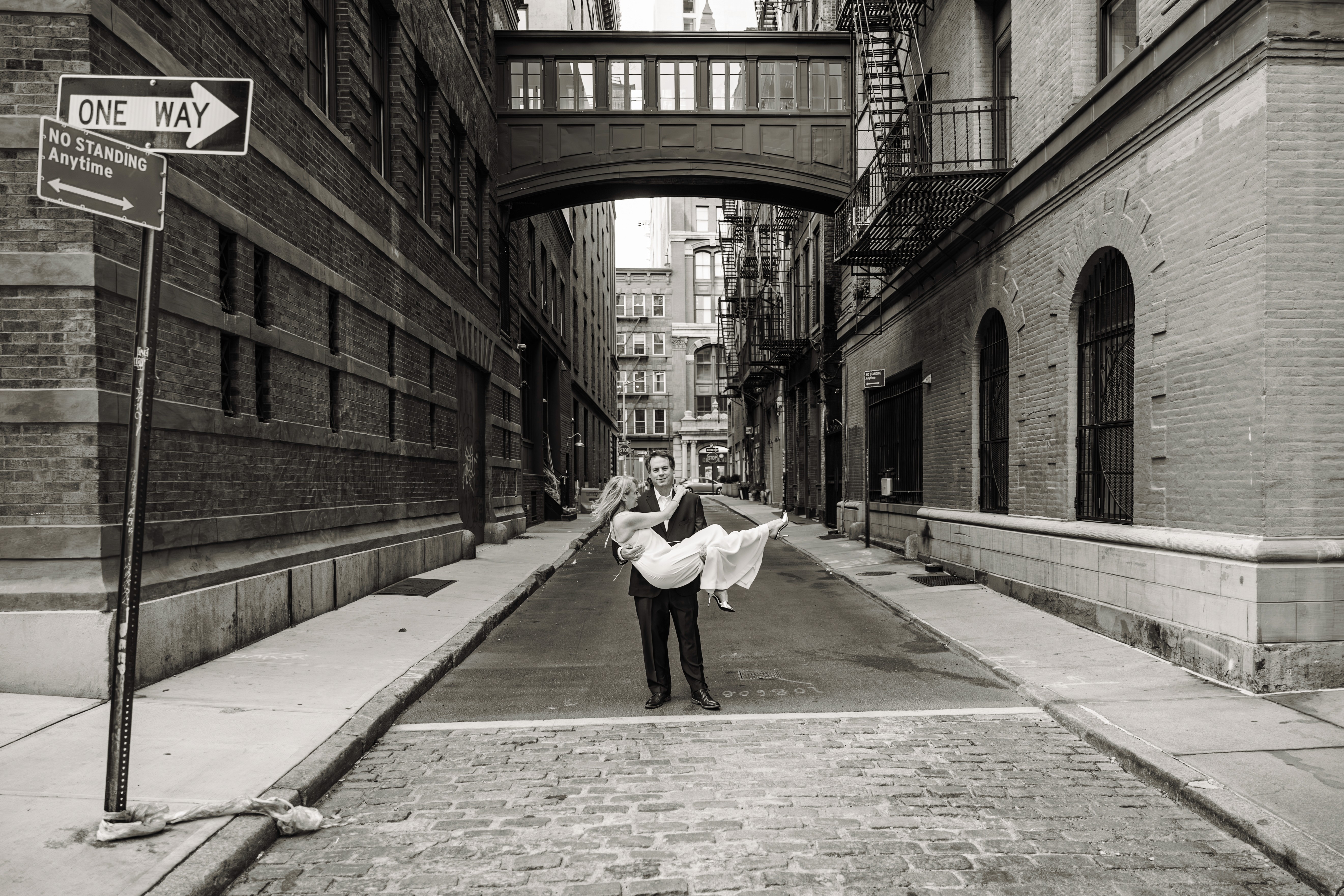 4:57 PMHim holding her like a baby, her laughing uncontrollably, him staring straight into the camera beneath the iconic Staple Street Bridge in Tribeca, NYC — cool, playful black and white couples photography by Lizz Spano Photography, New York City couples photographer.