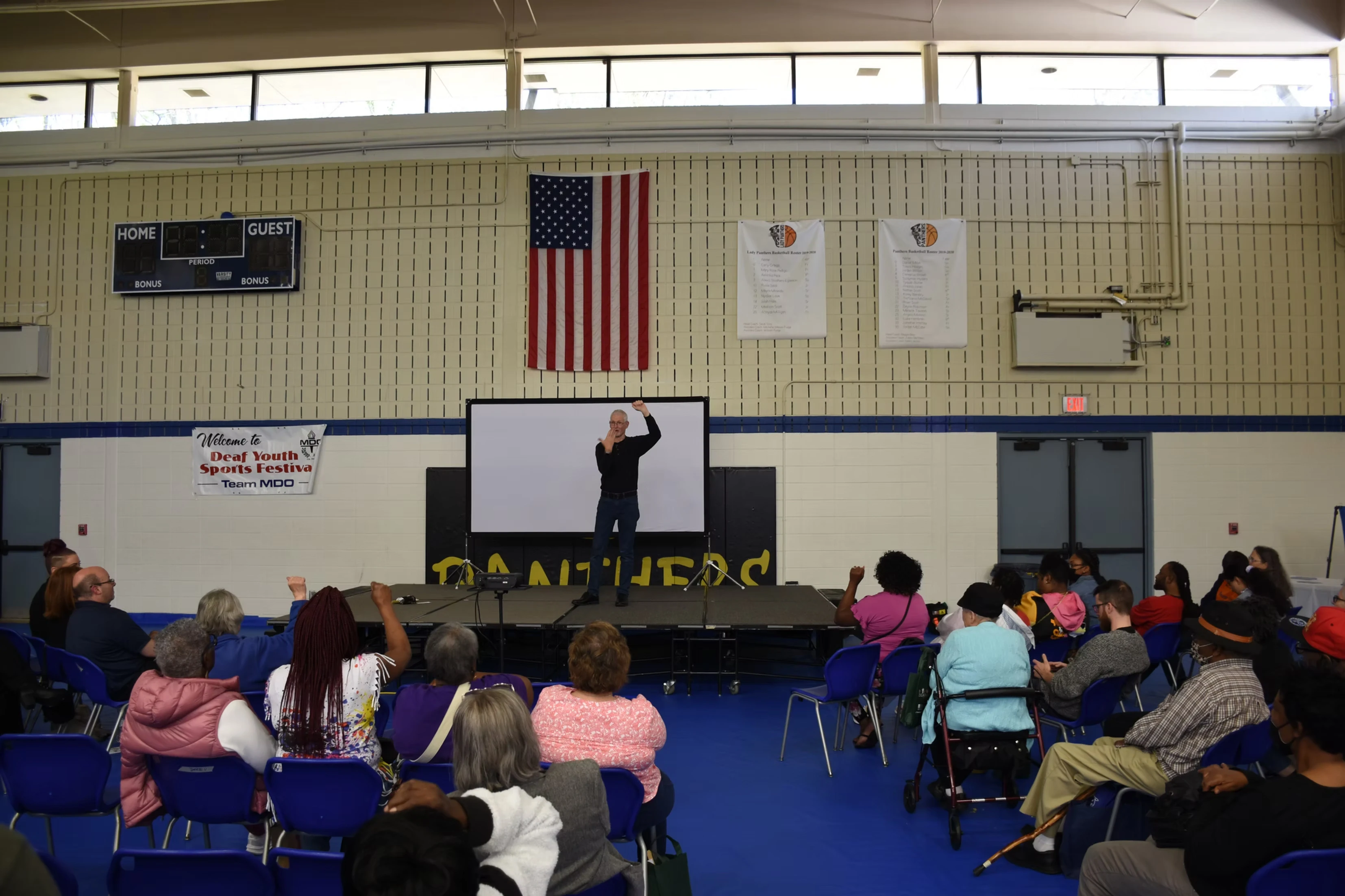 A man stands on a stage giving a presentation to a seated audience in a gymnasium.