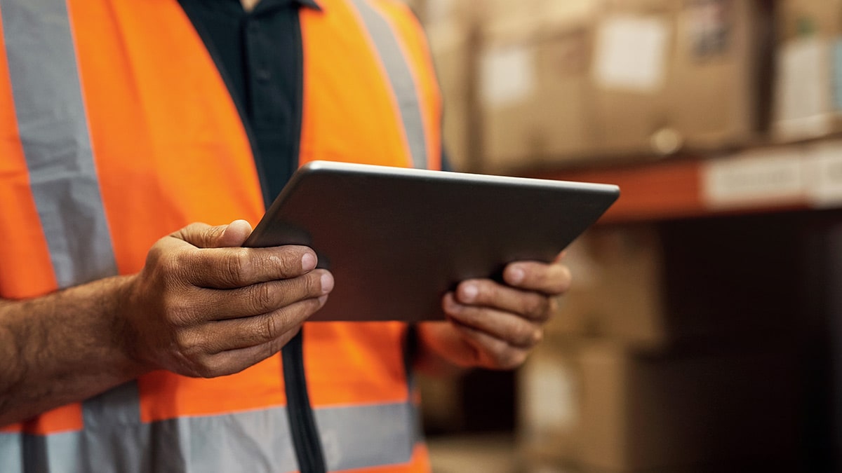 A person wearing a safety vest holds a tablet while standing in a warehouse with stacked boxes in the background.