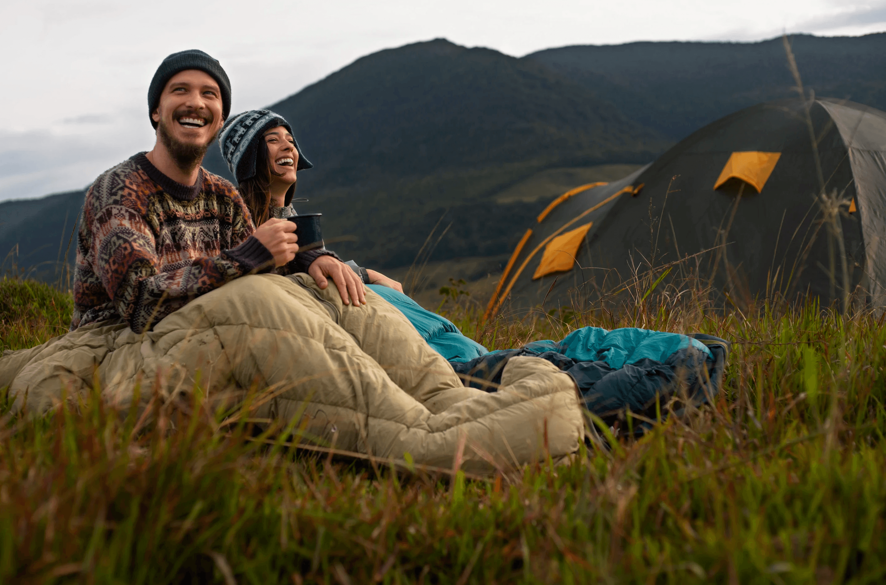 Two campers in sleeping bags by a tent in the mountains.