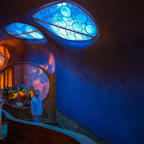 Persona de pie al pie de una escalera curva, debajo de ventanas de vidrio azul con patrones circulares, en una habitación con poca luz.