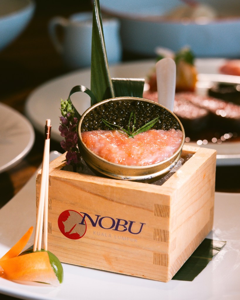 A beautifully presented Toro Tartare served in a wooden box with the Nobu Kuala Lumpur logo, accompanied by decorative elements like lime and carrot.