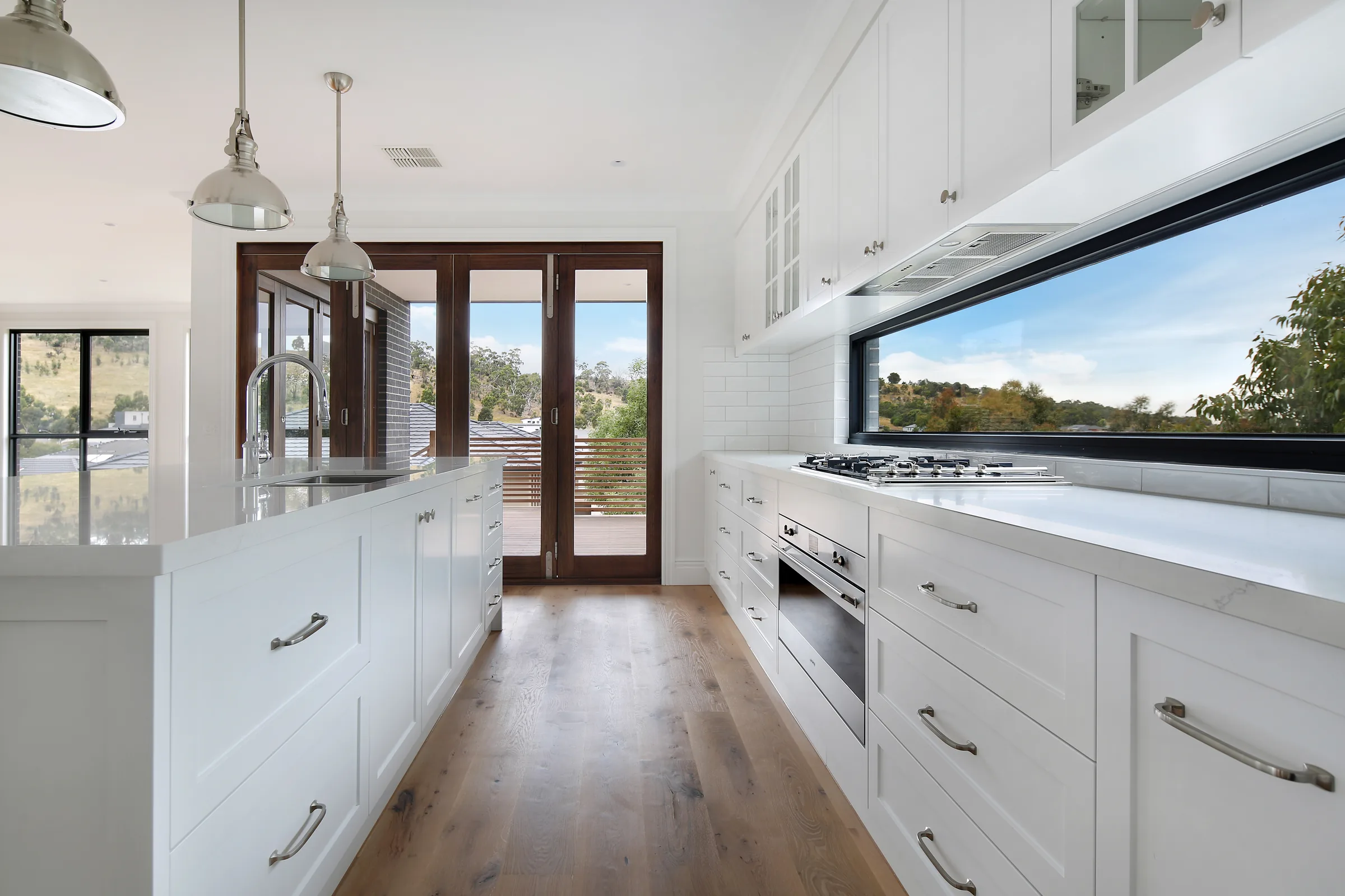 Galley kitchen with white shaker cabinetry, marble benchtops, polished-nickel dome pendants and a gas cooktop beneath a long black-framed window with a rural outlook, over oak floors with timber French doors to the deck