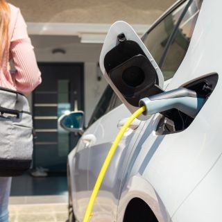Detailopname van een witte elektrische auto die wordt opgeladen bij een woning. De laadklep staat open en een felgele laadkabel is aangesloten. Op de achtergrond loopt een bewoner met een tas richting de moderne voordeur, wat de dagelijkse routine van thuisladen symboliseert.