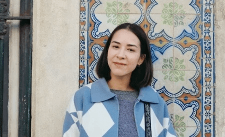 Caro smiling in a warm, outdoor setting with soft sunlight and a wall of painted Portuguese tiles in the background.