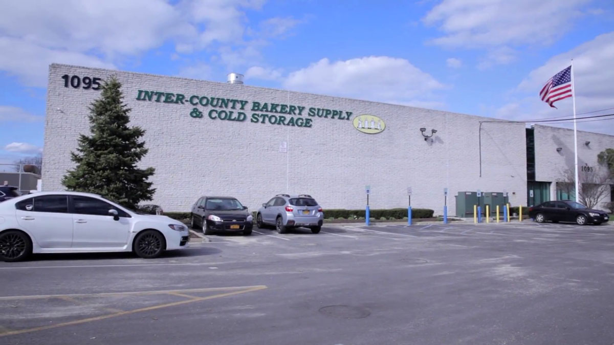 A large industrial building with the sign "Inter-County Bakery Supply & Cold Storage" is situated under a partly cloudy sky, with a U.S. flag fluttering nearby and a few parked cars in front.