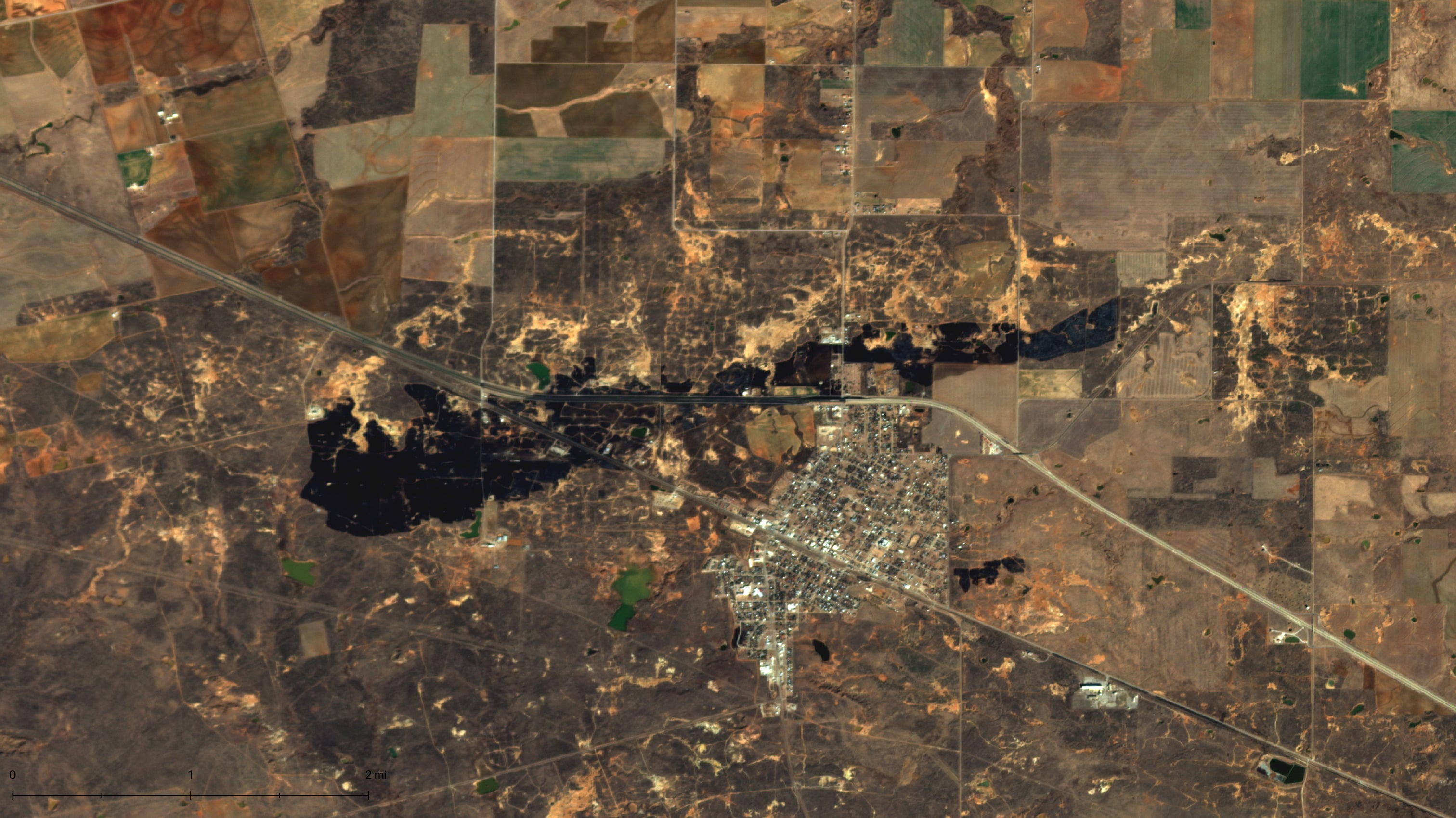 Aerial view of a town with a nearby body of water, surrounded by agricultural fields and patches of dry land, showcasing a mix of urban and rural landscapes.