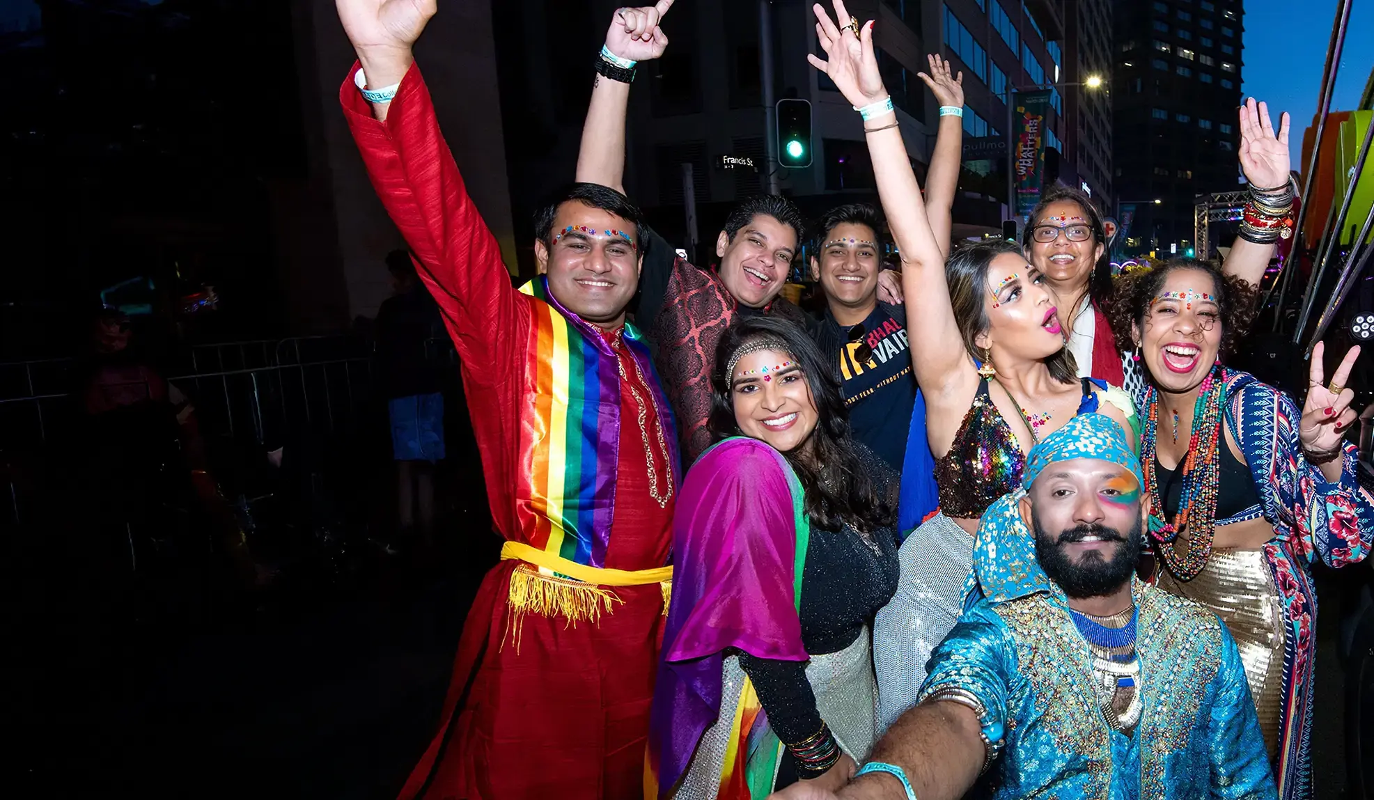 A group of people in colorful, festive outfits and face paint smile and pose with arms raised at a lively nighttime celebration or parade.