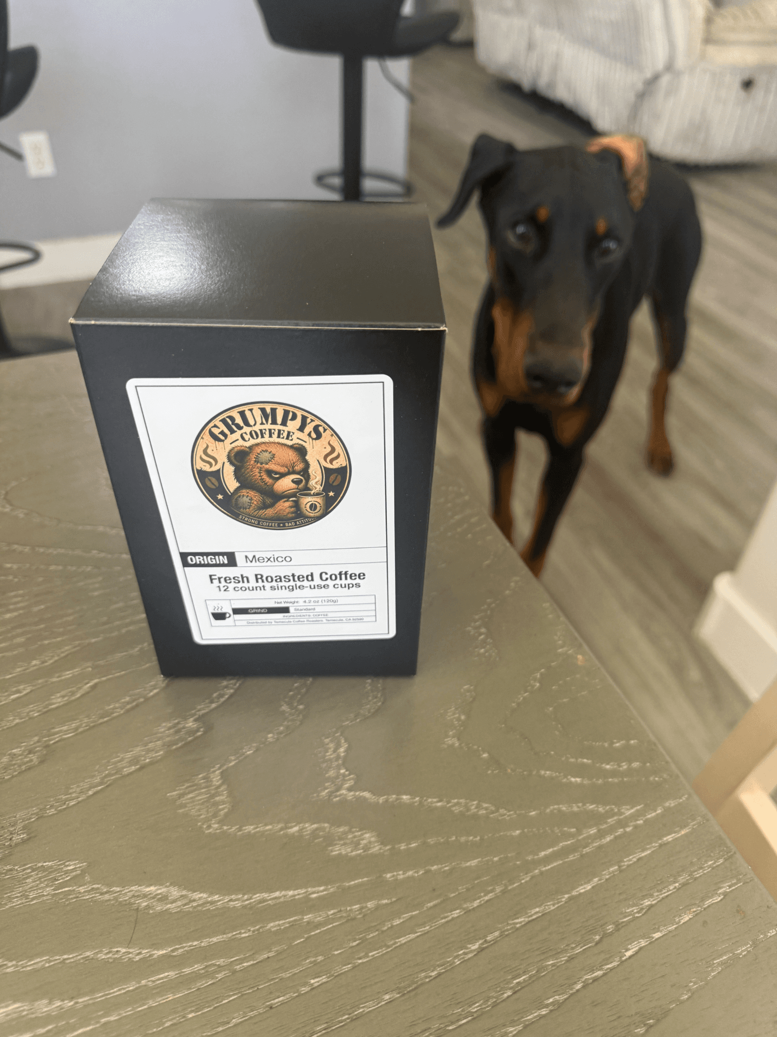 Grumpy’s Coffee single-use cup box on table with Doberman standing nearby indoors.