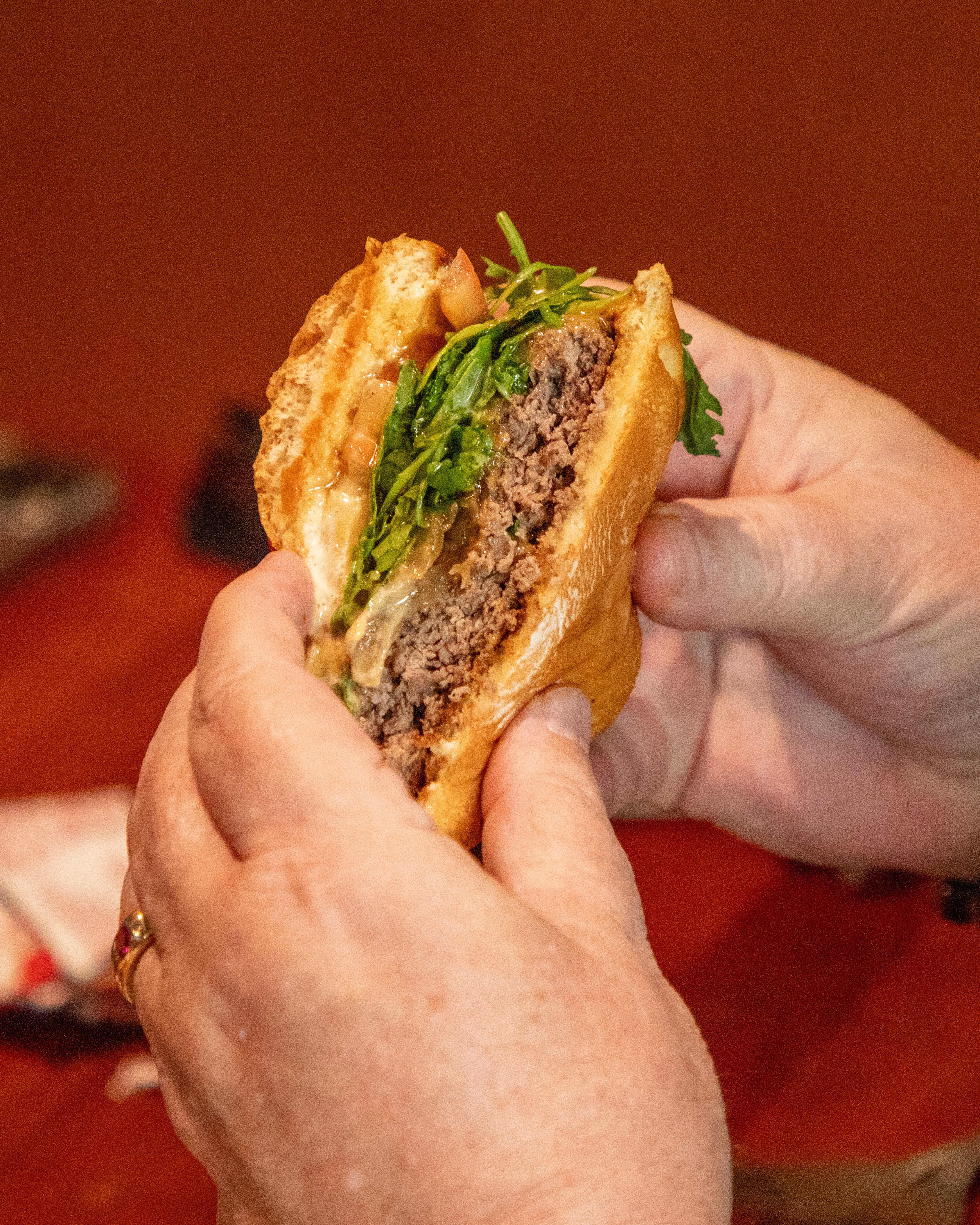 A close-up of a wok or large pan filled with golden noodles and vegetables, with a cook's hands adding more noodles, capturing steam and warm sunlight in a busy, rustic outdoor or market setting.