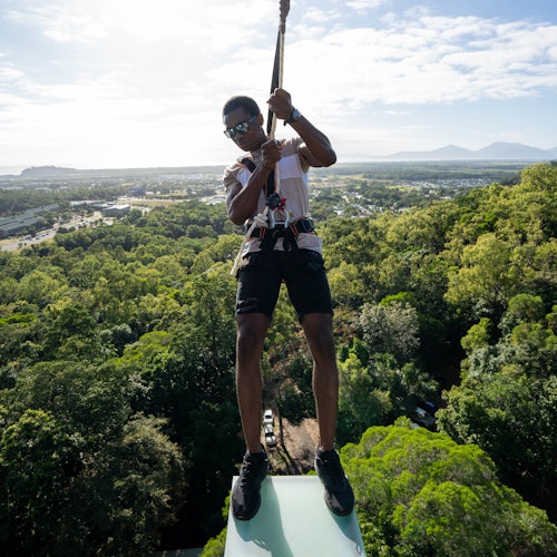 一个穿着安全带的人挂在森林上空的滑索上，背景中隐约可见城市景观和山脉。