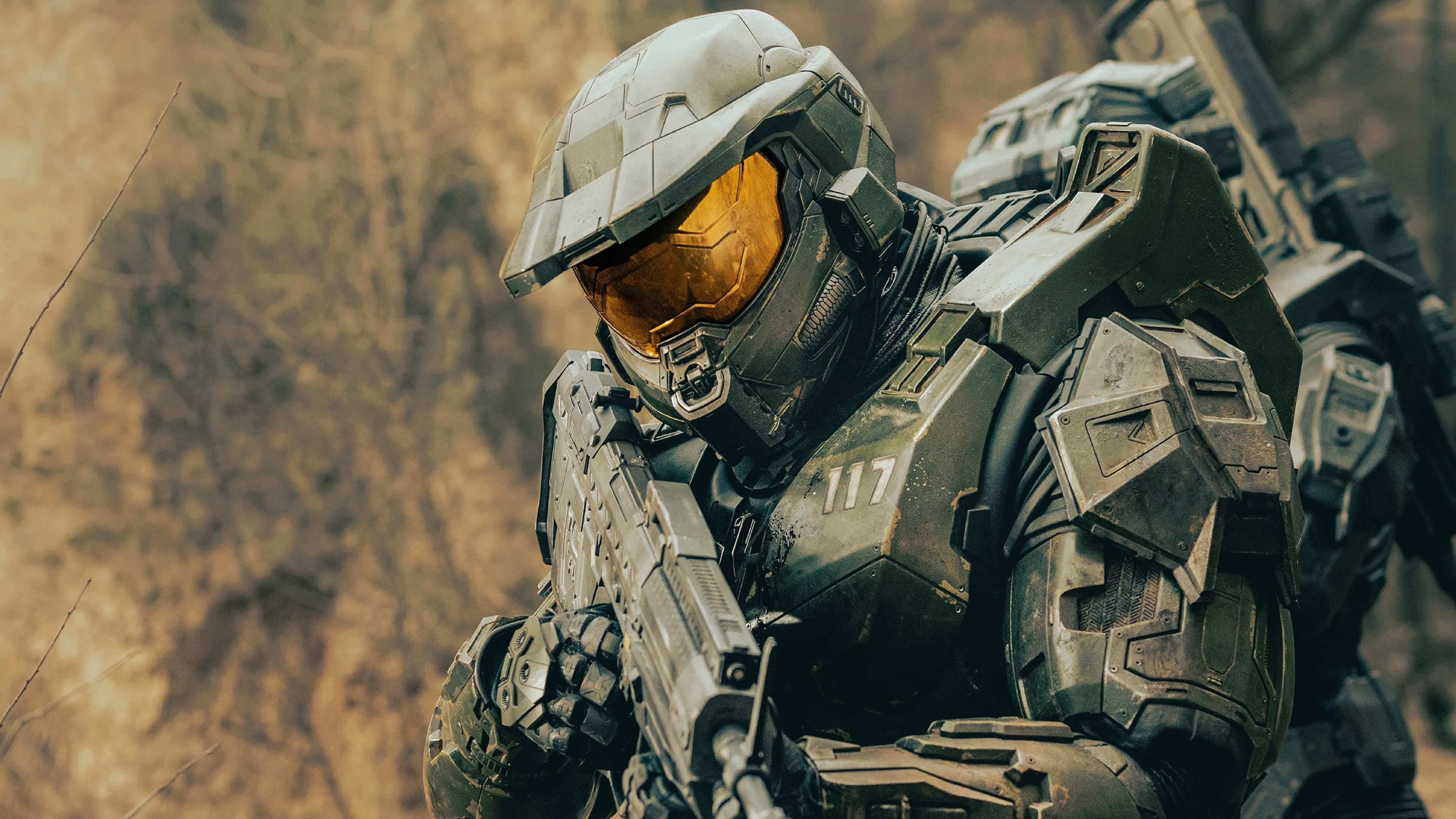 A futuristic soldier in advanced, metallic armour equipped with a reflective visor and holding a large weapon is poised in a forest environment, conveying intensity and focus.