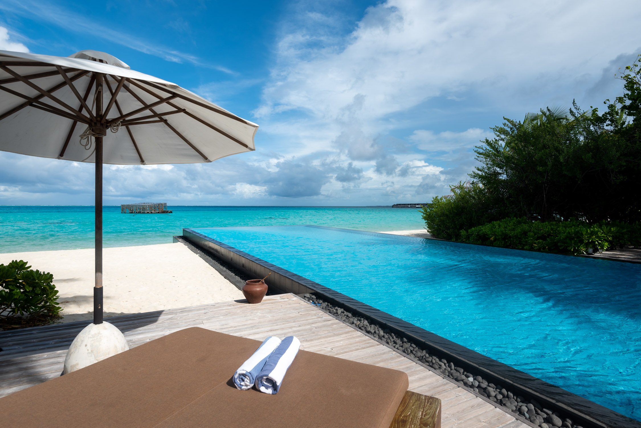 An infinity pool overlooks a tropical beach with white sand and turquoise water under a blue sky with white clouds.