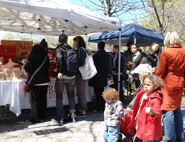 A white pop-up tent covers a table with food items, including what appears to be hot bread. Behind it, a blue pop-up tent is set up. Several people are gathered around the table and tents, including adults and children. A young child in a light-colored jacket and blue jeans stands near the table. A woman in a red coat is visible on the right side of the image. The scene is set outdoors on a paved area with trees in the background.