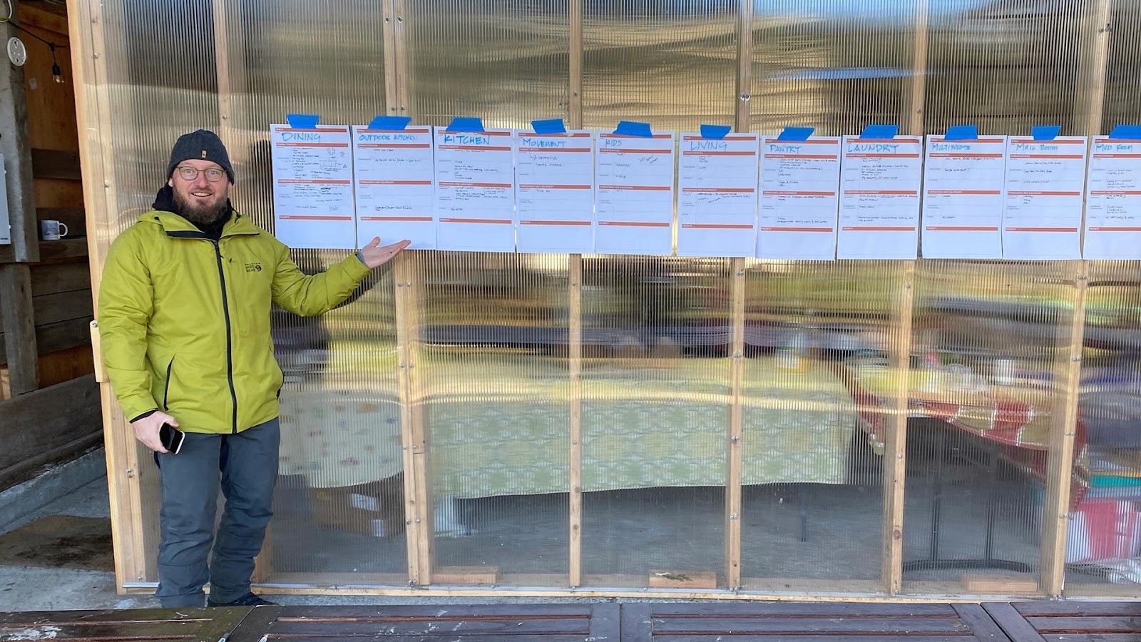 Bryan standing outside a greenhouse pointing to a row of posted project plans taped to the wall.