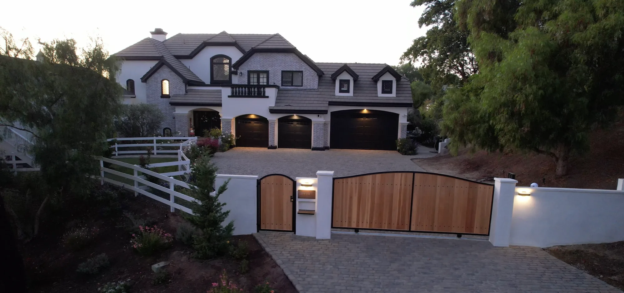 Gray paver driveway and timber entrance gates below a white two-story home at dusk.